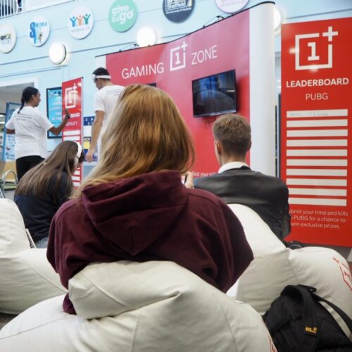 People at a gaming event sit on bean bags facing a screen with a Gaming Zone sign. A Leaderboard display for PUBG is nearby. The area is decorated with OnePlus branding and trophies are visible on a table. BAM Agency