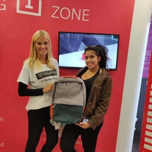 Two women smiling and holding a backpack in front of a red OnePlus display. One wears a branded t-shirt, and the other is in a brown jacket, holding a smartphone. BAM Agency
