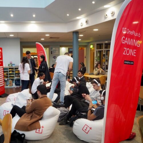 People are gathered in a gaming zone with red banners displaying OnePlus 6 Gaming Zone around them. Some individuals are sitting on white bean bags with headphones, using phones. Others are standing and interacting in the background. BAM Agency