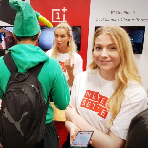 Two individuals wearing Never Settle t-shirts at a tech booth. One person faces forward, holding a phone and smiling. Another engages with a visitor wearing a green hat with a yellow beak. OnePlus 5 ad in the background. BAM Agency