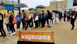 BAM Agency A diverse crowd stands outside, some clutching umbrellas, queued near a Wagamama sign. In this bustling urban scene, one person proudly holds a rainbow umbrella—an emblem of the youth market and the brands impact on vibrant city life. BAM Agency