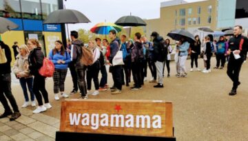 BAM Agency A diverse crowd stands outside, some clutching umbrellas, queued near a Wagamama sign. In this bustling urban scene, one person proudly holds a rainbow umbrella—an emblem of the youth market and the brands impact on vibrant city life. BAM Agency