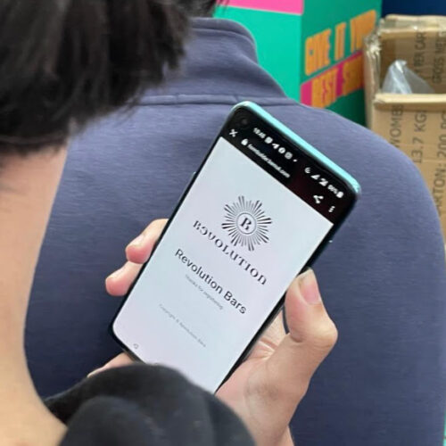 A person grasps a smartphone showcasing a webpage titled Revolution Bars. The screen is clearly visible, as the individual is slightly turned away. Colorful signs and a cardboard box enhance the lively backdrop, reminiscent of a buzzing night at Revolution. BAM Agency