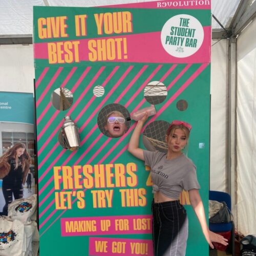A woman stands playfully beside a vibrant, carnival-style board with cut-out holes for heads and hands at Revolution Bars. The board reads Give It Your Best Shot and Freshers Lets Try This Again. Another person peeks through, smiling, capturing the lively nightlife spirit. BAM Agency