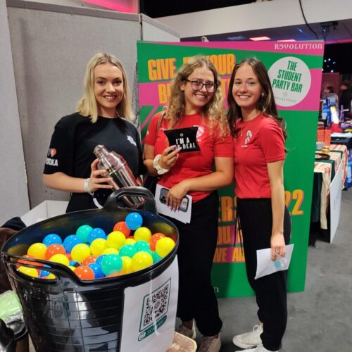 Three women stand at a booth with a container of colorful plastic balls. One holds a cocktail shaker, and two wear red shirts. A sign reads The Student Party Bar, reminiscent of Revolution Bars vibes, and flyers are held with a QR code visible in the foreground. BAM Agency
