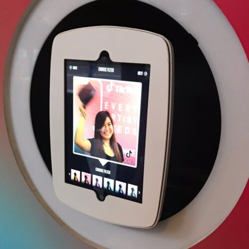 A tablet mounted on a circular frame displays a photo of someone smiling and waving a hat. The screen shows various filter options at the bottom. The background is blurred with a mix of pink and blue hues. BAM Agency