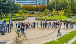 In a lively outdoor plaza, a large group forms a long, winding line near a 2019-themed food truck flanked by promotional flags. Surrounded by trees and a small pond, some relax on the grass while others stroll nearby. BAM Agency