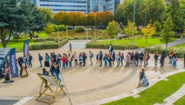 In a lively outdoor plaza, a large group forms a long, winding line near a 2019-themed food truck flanked by promotional flags. Surrounded by trees and a small pond, some relax on the grass while others stroll nearby. BAM Agency