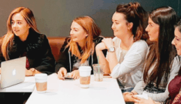 BAM Agency A group of seven women are sitting at a rectangular table, engrossed in a research project with a laptop and drinks. Light brown and gray walls create a warm backdrop as they discuss insights, some with coffee cups before them, contributing to the lively exchange on the youth market. BAM Agency