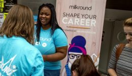 BAM Agency A woman in a blue shirt, embodying exceptional qualities, engages with people at a career event. Behind her is a banner that reads Milkround: Shape Your Career, featuring a graphic of a woman with glasses and bow tie. Attendees gather, interacting and holding papers. BAM Agency