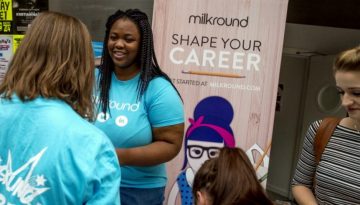 BAM Agency A woman in a blue shirt, embodying exceptional qualities, engages with people at a career event. Behind her is a banner that reads Milkround: Shape Your Career, featuring a graphic of a woman with glasses and bow tie. Attendees gather, interacting and holding papers. BAM Agency