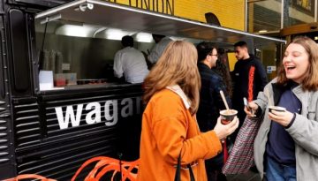 BAM Agency Two women happily chat while holding takeout containers in front of the award-winning food truck with wag visible. The open serving hatch buzzes with activity as other people line up, guided by their cravings for this experiential culinary delight. BAM Agency