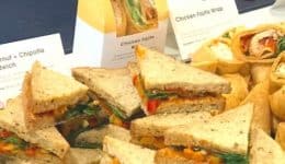 BAM Agency A platter of assorted sandwiches and wraps, featuring triangle-cut sandwiches with vibrant red and orange fillings, alongside verdant rolled wraps. The label proudly declares Honest Crust. Various signs provide details, showcasing a blend perfect for the refreshers crowd. BAM Agency