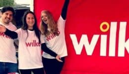 BAM Agency Four people smiling and posing next to a red Wilko banner outdoors. They are wearing white Wilko t-shirts and have their arms around each other, celebrating the 2018 National Freshers Tour. Grassy fields and trees provide a picturesque backdrop. BAM Agency
