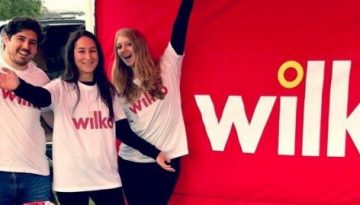 BAM Agency Four people smiling and posing next to a red Wilko banner outdoors. They are wearing white Wilko t-shirts and have their arms around each other, celebrating the 2018 National Freshers Tour. Grassy fields and trees provide a picturesque backdrop. BAM Agency
