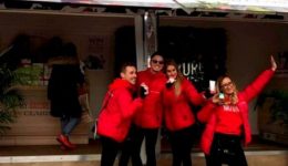 BAM Agency Three people in red jackets pose happily in front of a decorated mobile shop with open doors. The shop, part of a university tour, has white walls adorned with fruit and flower designs. Bystanders are visible nearby. BAM Agency