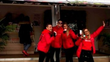 BAM Agency Three people in red jackets pose happily in front of a decorated mobile shop with open doors. The shop, part of a university tour, has white walls adorned with fruit and flower designs. Bystanders are visible nearby. BAM Agency