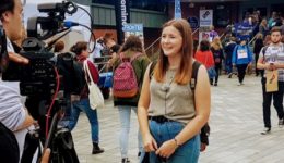 BAM Agency A young woman is being interviewed by a camera crew in an outdoor setting, highlighting the value of research. She stands among a bustling crowd, with people carrying backpacks. Stalls and balloons dot the lively background, capturing the essence of vibrant market research in action. BAM Agency