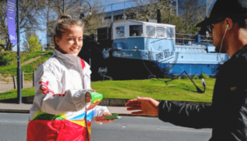 BAM Agency A child is handing a small item to an adult wearing a black cap and backpack on a sunny street, as part of the Just Eat campaign. A blue boat is in the background, surrounded by trees, while red JUST EAT text decorates the left—a perfect Whopper Wednesday scene. BAM Agency