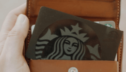 BAM Agency A hand holds an open brown leather wallet, revealing a card with a black and white logo featuring a crowned mermaid, hinting at exclusive loyalty schemes. The wallets interior contrasts against a blurred backdrop, appealing to the savvy youth market. BAM Agency