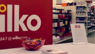 BAM Agency Inside the Burnley Wilko store, a large red Wilko sign adorns the left side. A white table showcases a bowl of colorful wrapped candies, papers, and a sign, while shelves brimming with various products stand ready in the background. BAM Agency