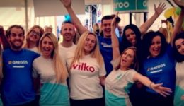 BAM Agency A group of smiling promo staff, proudly wearing branded shirts from Wilko, Argos, and other companies, cheerfully pose with their arms raised. They are in a bright indoor setting decorated for marketing success with colorful banners and balloons. BAM Agency