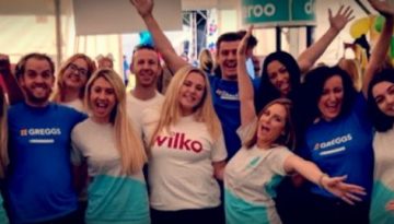 BAM Agency A group of smiling promo staff, proudly wearing branded shirts from Wilko, Argos, and other companies, cheerfully pose with their arms raised. They are in a bright indoor setting decorated for marketing success with colorful banners and balloons. BAM Agency