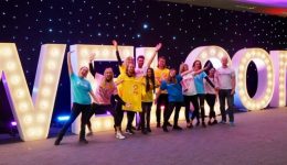 BAM Agency A group of smiling people wearing colorful shirts stand in front of large, illuminated letters spelling WELCOME. Celebrating exciting news, they pose with arms raised in a festive indoor setting, ready to embrace the freshers season. BAM Agency