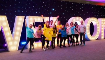 BAM Agency A group of smiling people wearing colorful shirts stand in front of large, illuminated letters spelling WELCOME. Celebrating exciting news, they pose with arms raised in a festive indoor setting, ready to embrace the freshers season. BAM Agency