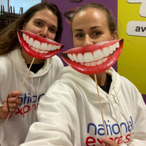 BAM Agency Two people in white hoodies, with a colorful National Express logo, hold oversized smiling mouth cutouts on sticks in front of their faces. They stand against a backdrop of vibrant colors and partial text, embodying the joy of every transport service journey. BAM Agency