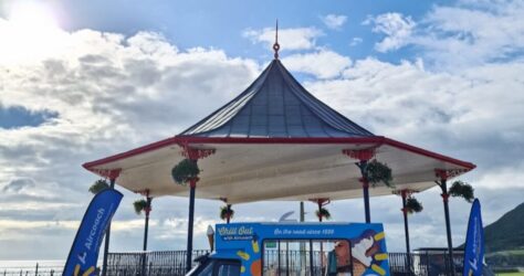 A blue and white ice cream van is parked in front of a seaside bandstand with hanging flower baskets. Two matching flags stand on either side. The sky is bright with scattered clouds and sunlight. BAM Agency