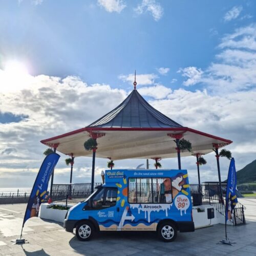 A blue and white ice cream van is parked in front of a seaside bandstand with hanging flower baskets. Two matching flags stand on either side. The sky is bright with scattered clouds and sunlight. BAM Agency