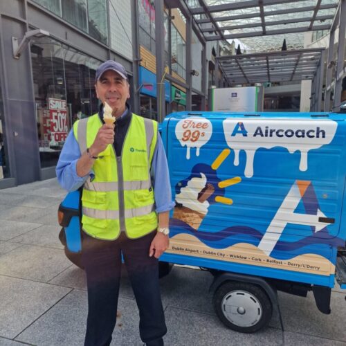A man in a yellow safety vest smiles whilst holding an ice cream cone, standing next to a small blue Aircoach trolley offering free 99s, on a city street with shops in the background. BAM Agency