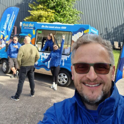 A smiling man in a blue jacket takes a selfie in front of an ice cream van with “Chill Out with Aircoach” branding, while three other people in blue jackets and one person in a tan jacket stand nearby. BAM Agency
