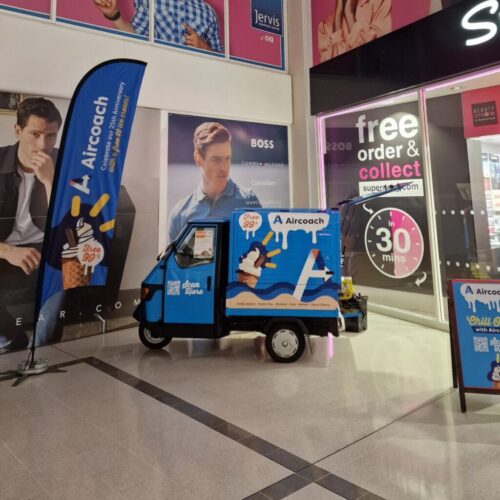 A small blue Aircoach promotional vehicle with banners is parked indoors near shopfronts, including a free order & collect shop and a large BOSS advertisement featuring a man in a suit. BAM Agency