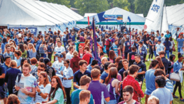 BAM Agency A large crowd gathers at the outdoor Freshers campaign under multiple white tents. People in colorful shirts hold signs, creating a lively setting with greenery and a banner enhancing the scenes vibrancy. BAM Agency