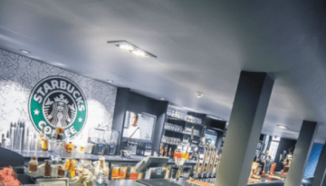 BAM Agency A modern bar with a stylish interior features a curved counter, stools, and various brands of bottles on display. The ceiling has track lighting, and a large circular logo is visible on the wall. Theres a window on the right with sunlight streaming in. BAM Agency