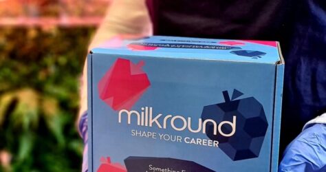 A person wearing a dark apron and gloves holds a blue box with colorful graphics and the text milkround - SHAPE YOUR CAREER. The background is blurred, with a neon-like sign partially visible. BAM Agency