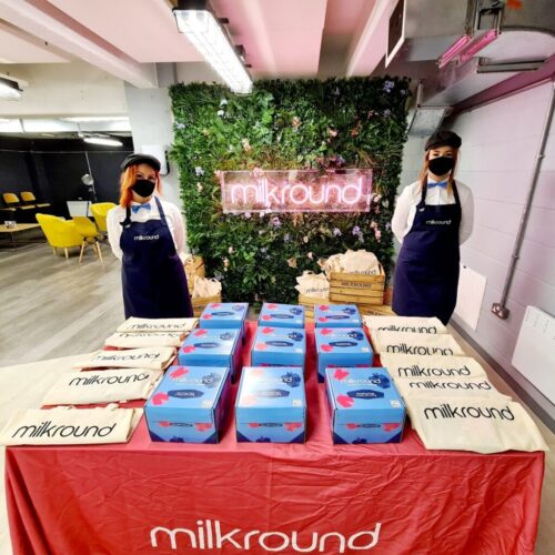 Two people wearing black masks and navy aprons stand behind a table with multiple blue boxes and branded tote bags. A greenery wall with a neon sign reading milkround is in the background. BAM Agency