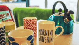 BAM Agency Five colorful mugs and a teal kettle are arranged on a wooden table. Each mug boasts a unique pattern or text, while an advertising agents retro-style plaque is subtly visible in the blurred background beside a chair with towels. BAM Agency