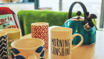 BAM Agency Five colorful mugs and a teal kettle are arranged on a wooden table. Each mug boasts a unique pattern or text, while an advertising agents retro-style plaque is subtly visible in the blurred background beside a chair with towels. BAM Agency