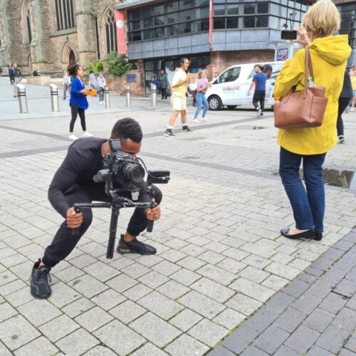 A man in black squats whilst filming with a professional camera on a city street, as a woman in a yellow jacket stands nearby taking a photo with her mobile. People walk and historic buildings are visible in the background. BAM Agency