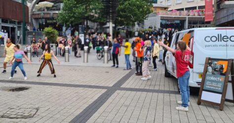 A lively outdoor scene in a city square shows dancers performing for a gathered crowd. People watch from the pavement, while a person in red stands near a CollectPlus sign and van. Shops and trees are visible in the background. BAM Agency
