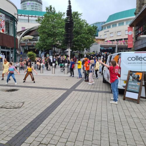 A lively outdoor scene in a city square shows dancers performing for a gathered crowd. People watch from the pavement, while a person in red stands near a CollectPlus sign and van. Shops and trees are visible in the background. BAM Agency