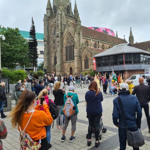 A crowd gathers near a historic stone church with a tall spire. People watch a street performance or demonstration, some filming with mobiles. The sky is cloudy and buildings surround the area. BAM Agency