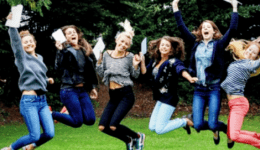 BAM Agency A group of students outdoors, joyfully jumping in the air with trees in the background. They hold white papers and are dressed casually in jeans and jackets, smiling widely. BAM Agency