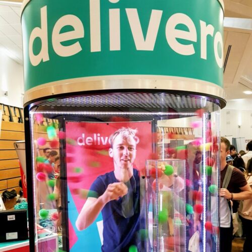A person is inside a clear booth with colorful balls swirling around. The booth has the word delivero on top. The person is smiling and reaching for the balls. A crowd is visible in the background. BAM Agency