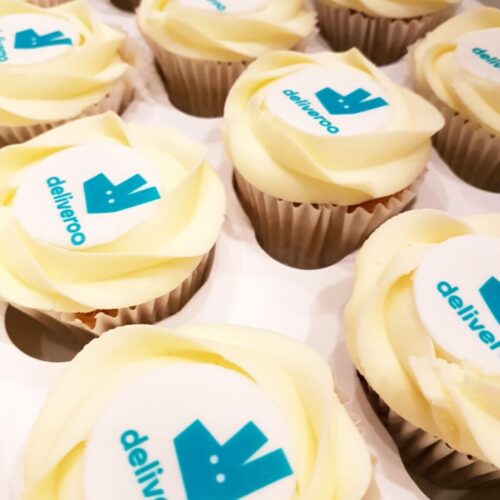 A tray of cupcakes with creamy frosting, each topped with an edible decoration displaying the Deliveroo logo, featuring a turquoise kangaroo image and the word Deliveroo in matching color. BAM Agency