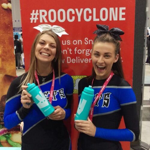 Two cheerleaders, wearing blue and black uniforms with bows in their hair, smile while holding teal bottles. They stand in front of a red backdrop with the hashtag ROOCYCLONE. BAM Agency
