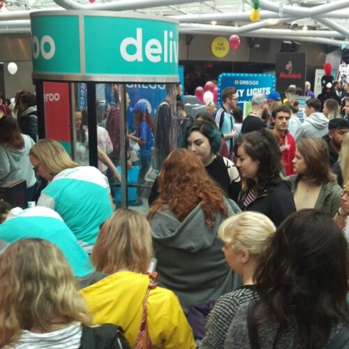 A crowded indoor event with many people gathered around various booths. Visible brand logos include Deliveroo, Greggs, and Nandos. Attendees are engaged and exploring different stalls under a skylit ceiling. BAM Agency